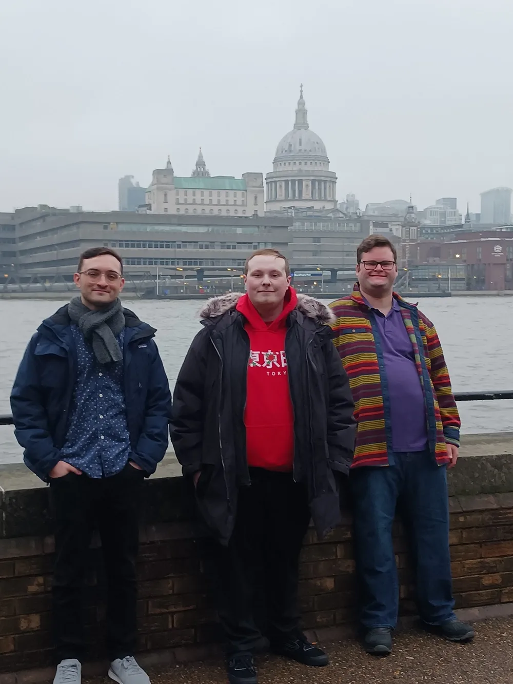 Three young men in winter clothing stand on the banks of the Thames River in central London with St. Paul's cathedral in the background.