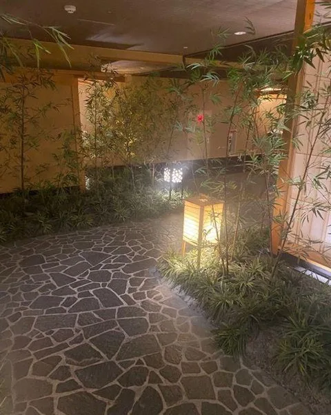 A picture of a stone road leading through some thin bamboo sticks and paper lanterns. Brown screens are in the background.