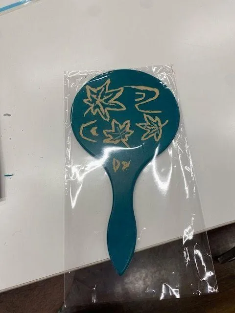 A blue mirror is shown in the photo with the back adorned with wooden carvings resembling leaves blowing in the wind. The initials “D.Y” are inscribed below the carvings.