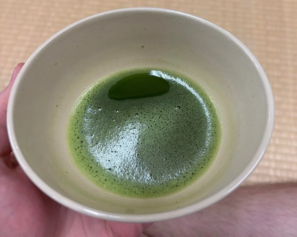 A hand holds a ceramic bowl with freshly whisked matcha green tea, resting over a traditional tatami mat floor.
