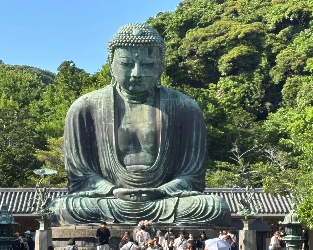 A large bronze statue of the Great Buddha (Daibutsu) sits peacefully in a meditative pose, surrounded by lush green trees and visitors at its base under a clear blue sky.