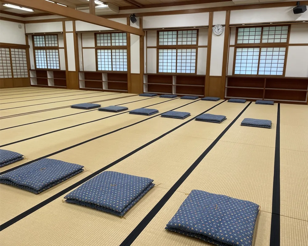 On a floor made of tatami, flat square pillows are arranged in three spaced-out rows, with six pillows per row. The room has many wood beams supporting it, and has paper sliding windows on the far wall.