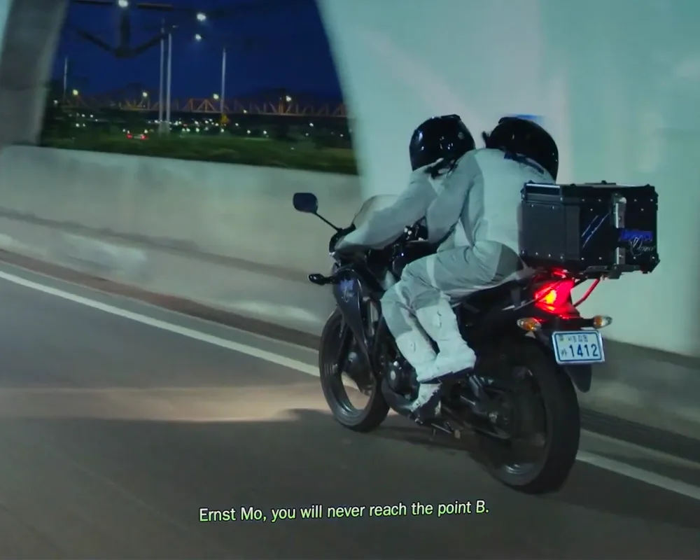 Two women driving on a motorcycle through a road with the subtitles “Mo, you will never reach the point B.”