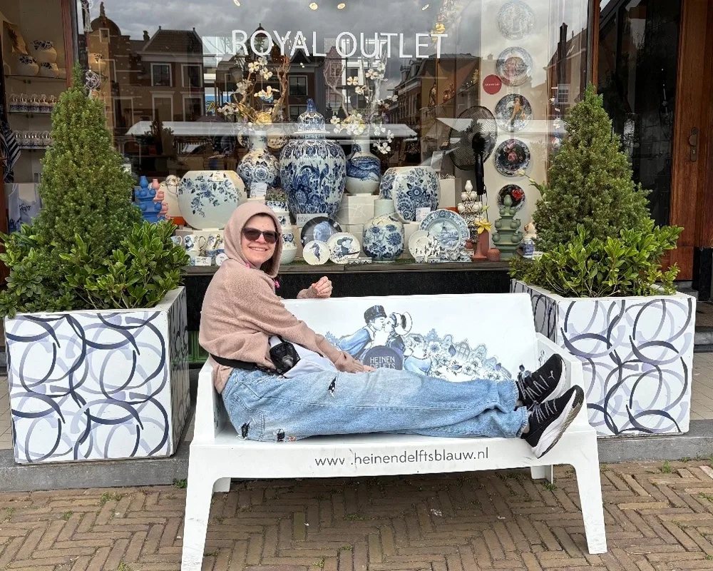 Photo of smiling woman in jeans and hoody sitting lengthwise on a bench in front of a storefront called the Royal Outlet. Dominant color is blue.