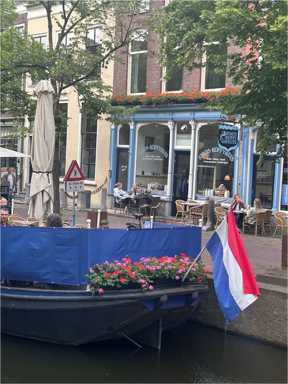 photo of coffee house with blue store front. People sitting at cafe tables outside along the canal. Boat transom with flowers and Netherlands flag visible in forefront.