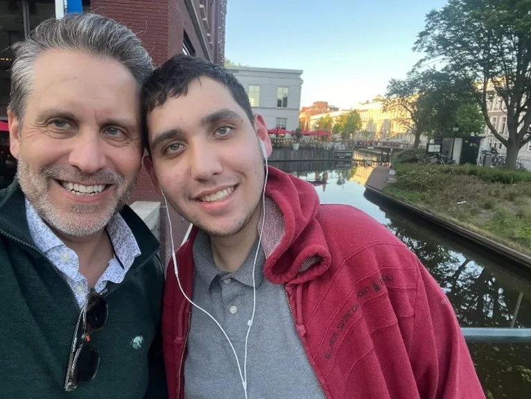 A man and his 21-year-old son stand with their heads together on a bridge over a canal