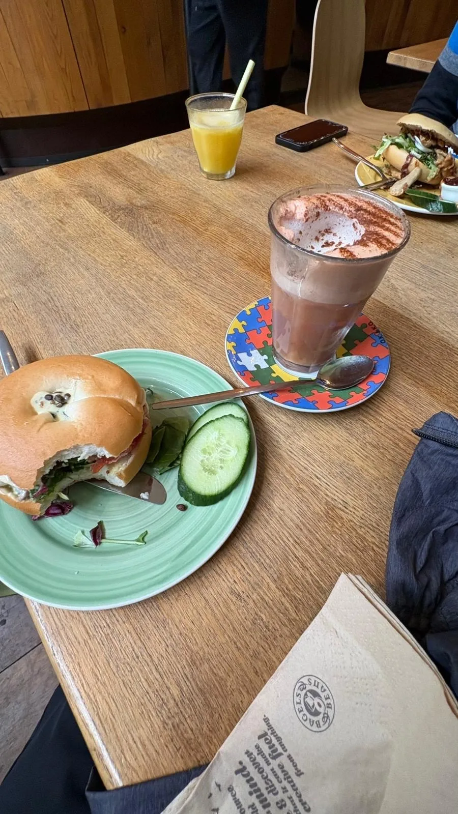 Formica table top with bagle sandwhich with a bite taken out accompanied by a chocolate shake and another plate and juice drink in the background.