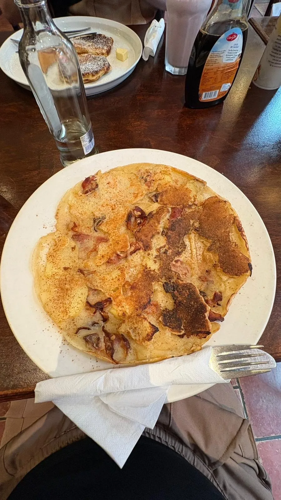 Image of a large pancake on a white place with glass bottle of water and syrup bottle next to it on the table.