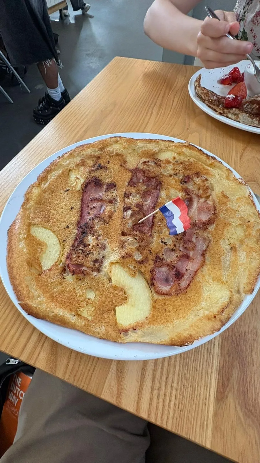 On a table a dish bearing a plate sized pancake adorned with applesclices and speared with a small Netherlands flag toothpick