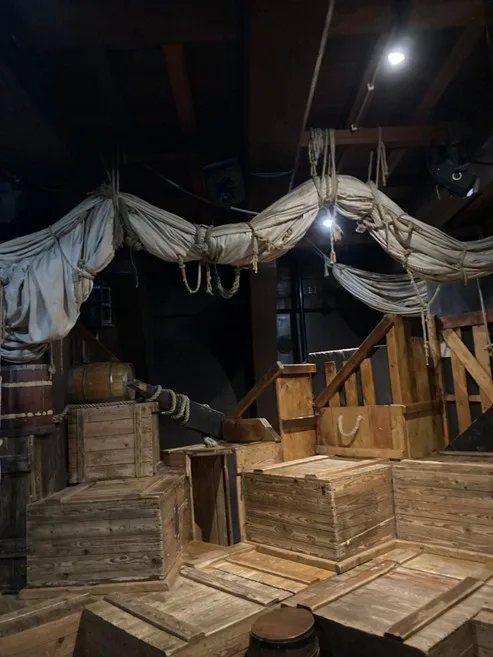 Image of a cargo hold of a wooden ship. The room is lit by artificial light and filled with wooden crates and barrels. A spare canvas sail is furled and stored hanging in slings along the upper beams.