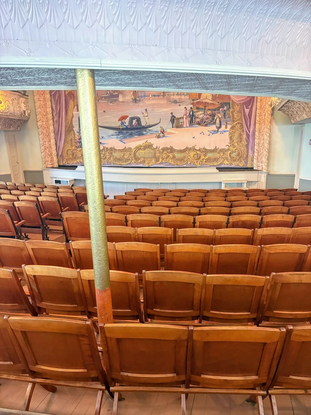 View of stage with tapestry in background from behind many rows of wooden seats
