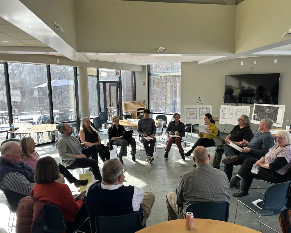 A group of 15 people sitting in a circle and having a conversation. Behind them are three easels with floor plans of buildings and aerial photography of campus