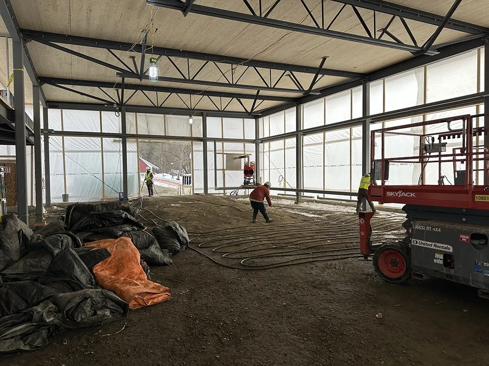 Interior of building under construction with worker laying out heating coils on the ground.