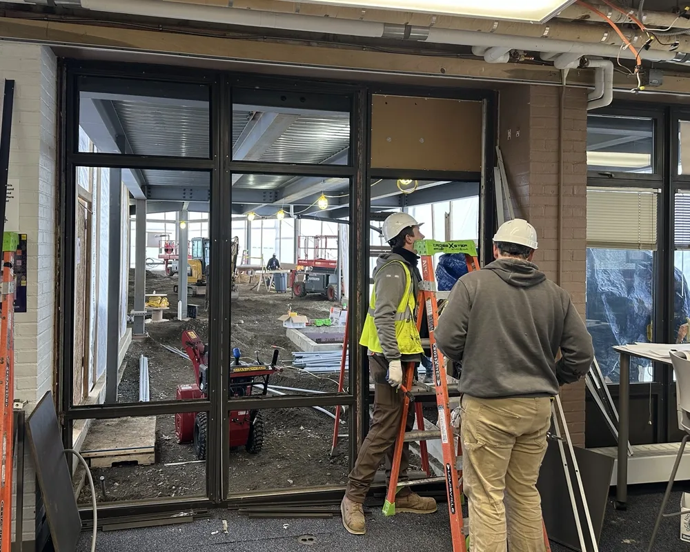Two constructions workers near ladder in front of large window frame looking into large open space