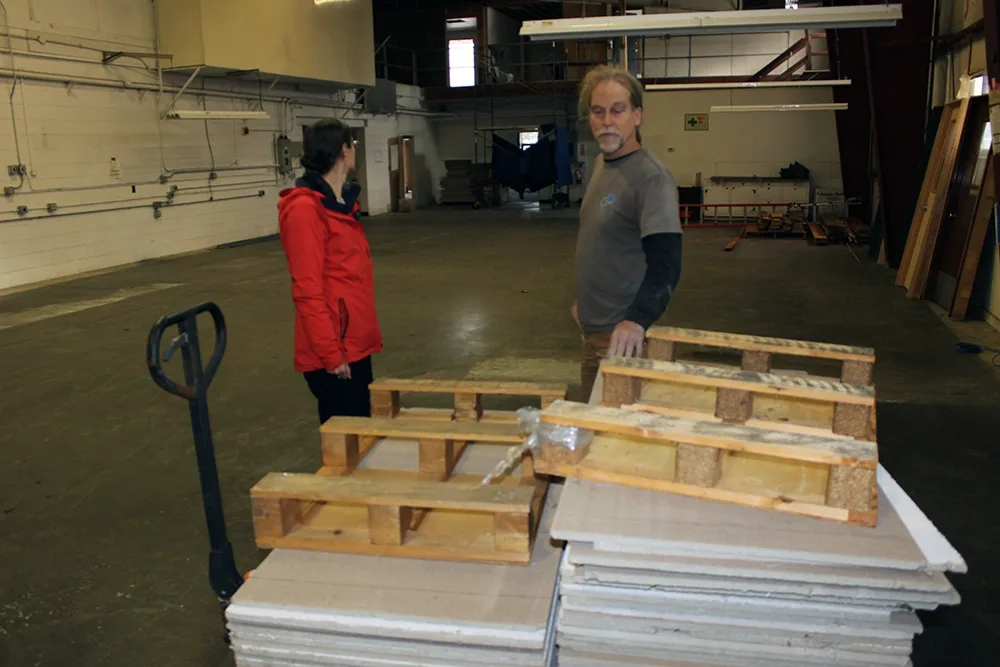 gray haired man and woman in red jacket standing next to a stack of ceiling tiles in a large warehouse space.