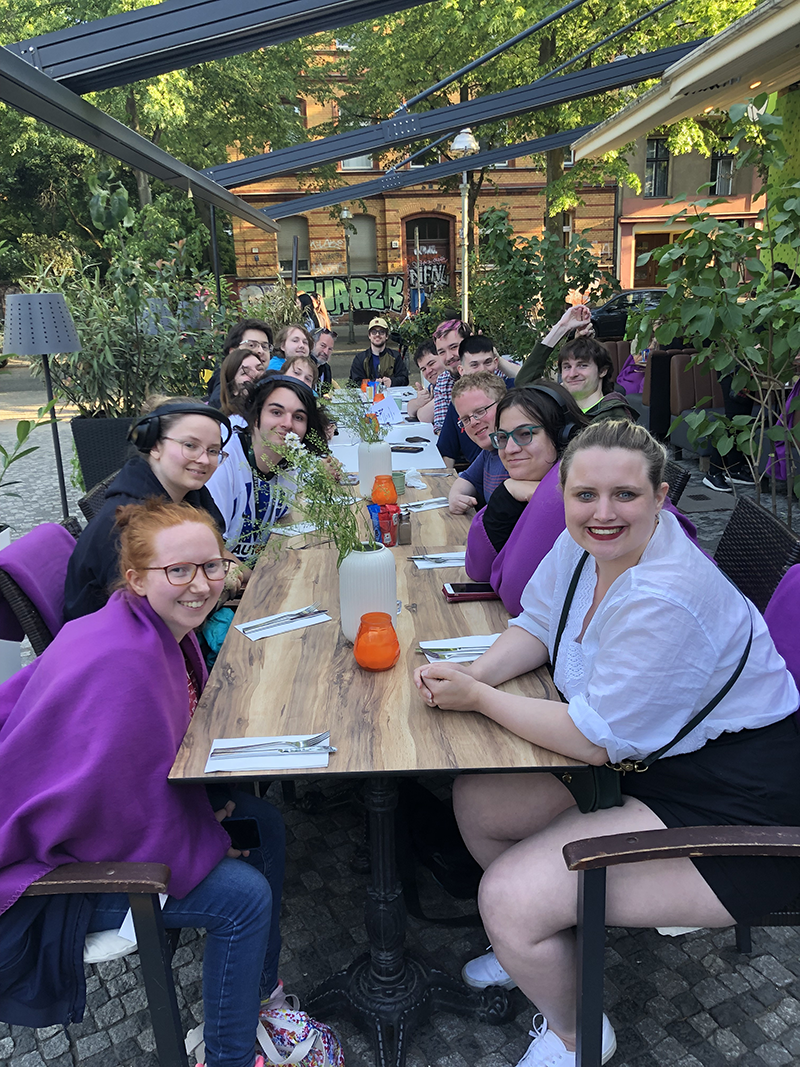 A group of smiling students sitting at a long wooden table on outside, waiting to be served dinner at a restaurant