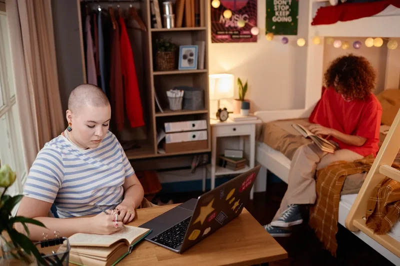 View of a dorm room with a student taking notes in front of a laptop with another student in a red shirt sitting on a bunk bed reading a book.