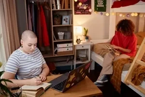 View of a dorm room with a student taking notes in front of a laptop with another student in a red shirt sitting on a bunk bed reading a book.