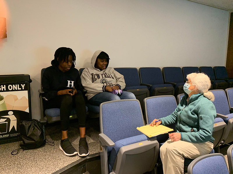A woman with white hair and protective face mask turns around in her chair to talk with two female students sitting behind her.