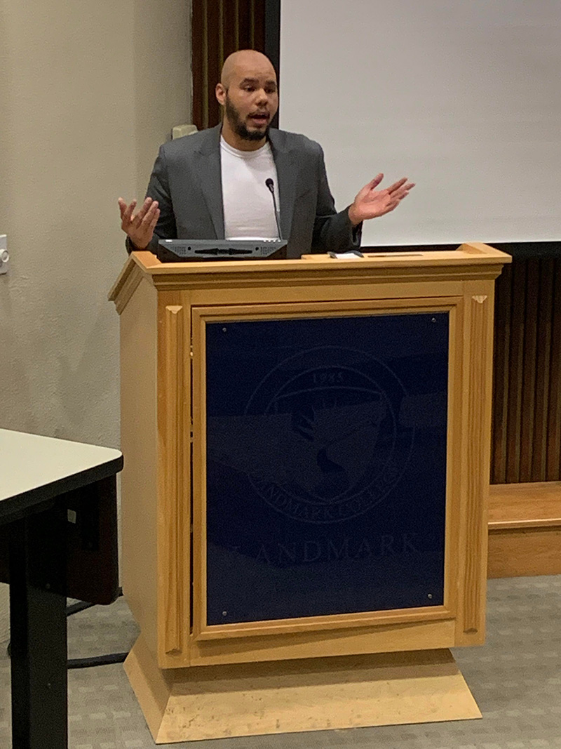 Audience view of Steffen Gillom at podium. He has a shaved head and beard and is wearing a gray blazer over a white t-shirt.