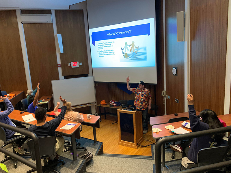 Audience view of student in bright patterned shirt making a presentation. He is raising his hands and other audience members also have their hands raised.