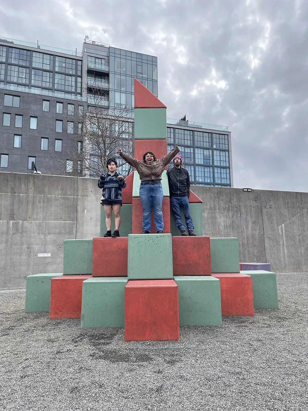 3 students standing on colored blocks in an outdoor sculpture park. The student in the center has her arms raised above her head in celebration