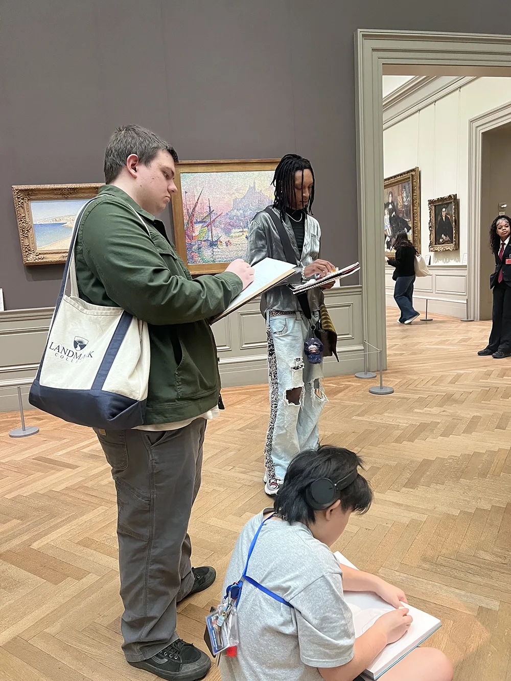 3 students in museum gallery space sketching on drawing pads. Two are standing and one is sitting on the floor.