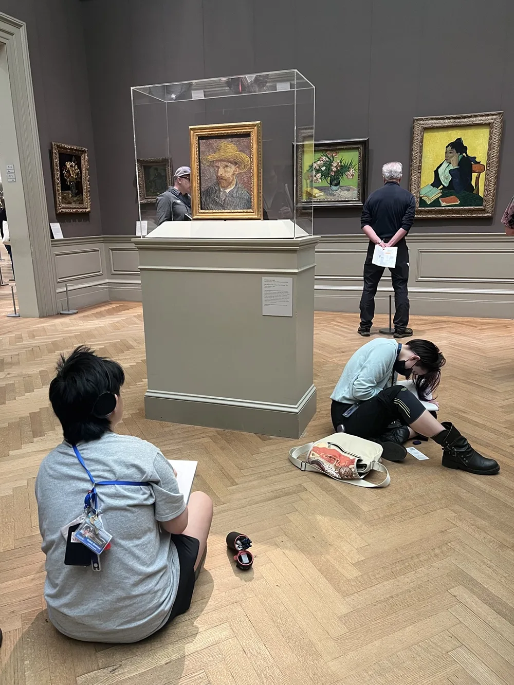 Two students sketching while seated on the floor a museum gallery. One has their back to a large case containing a painting in the center of the room. The other student is facing that case.