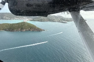 a view from an airplane window, with the underside of the plane’s wing above, looking out over tropical islands with ships in the bay and white buildings scattered in the hills.