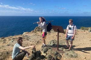 a student on the left builds a cairn of rocks; a second, in the center, holds a long stick over his shoulder; a third, on the right, reads an informational panel