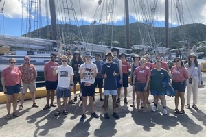 A photo of 18 people standing on a concrete dock in front of a sailing ship with masts, sails, and rigging, with tropical hills in the background.