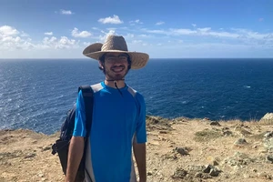 a smiling student wearing a sun hat on a hill with the sea behind him