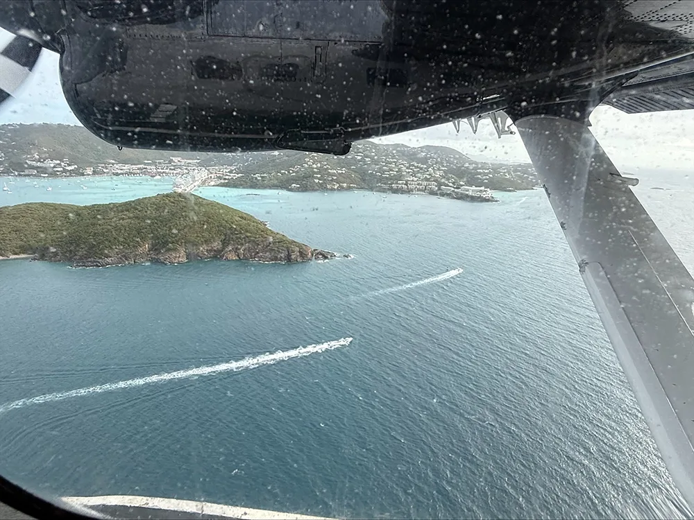 a view from an airplane window, with the underside of the plane’s wing above, looking out over tropical islands with ships in the bay and white buildings scattered in the hills.