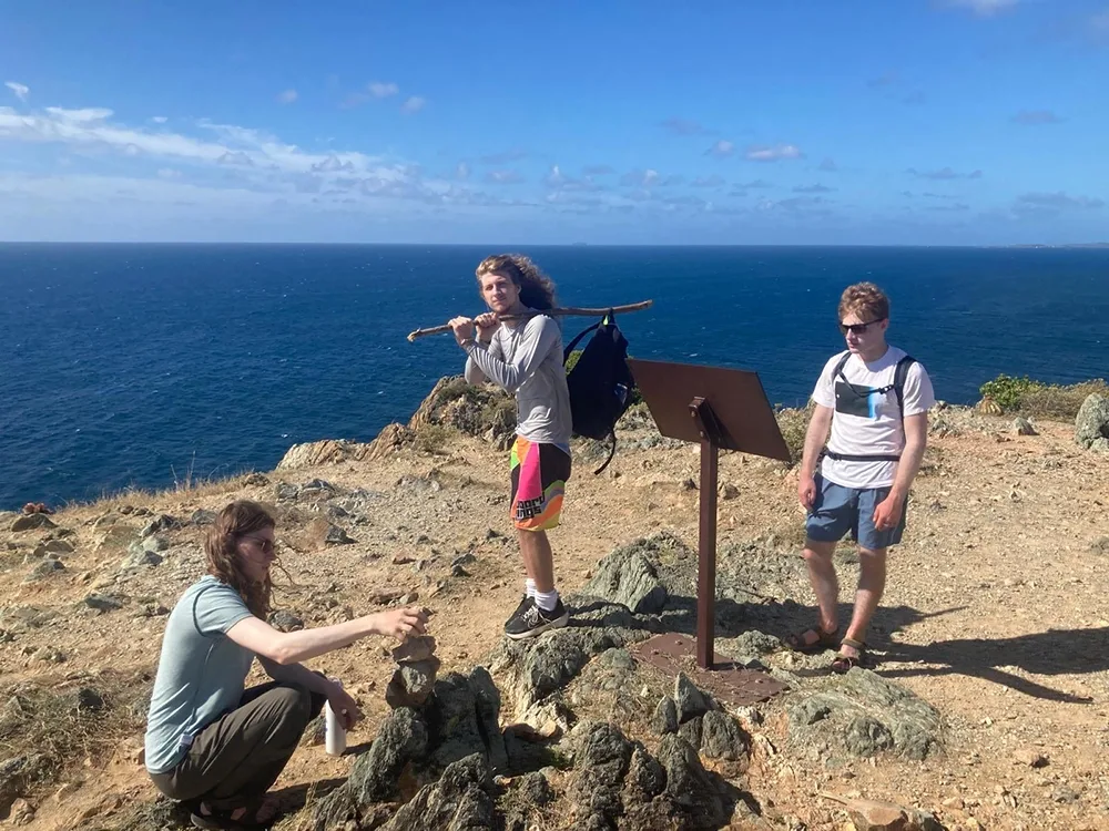 a student on the left builds a cairn of rocks; a second, in the center, holds a long stick over his shoulder; a third, on the right, reads an informational panel