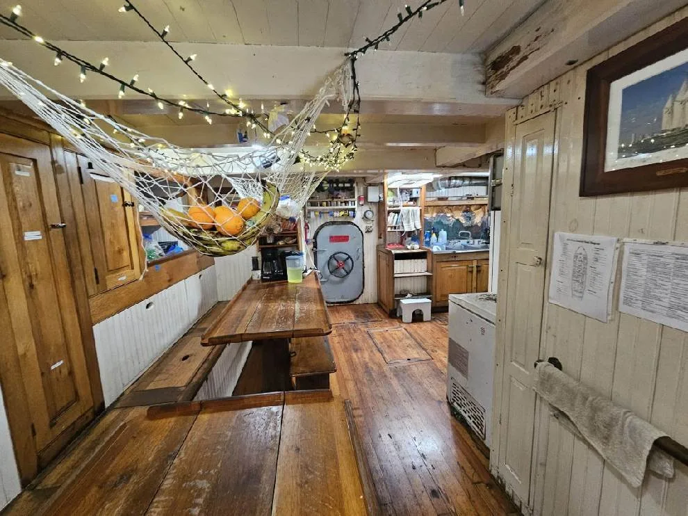 kitchen area on ship with long wooden table, refrigerator and sink.