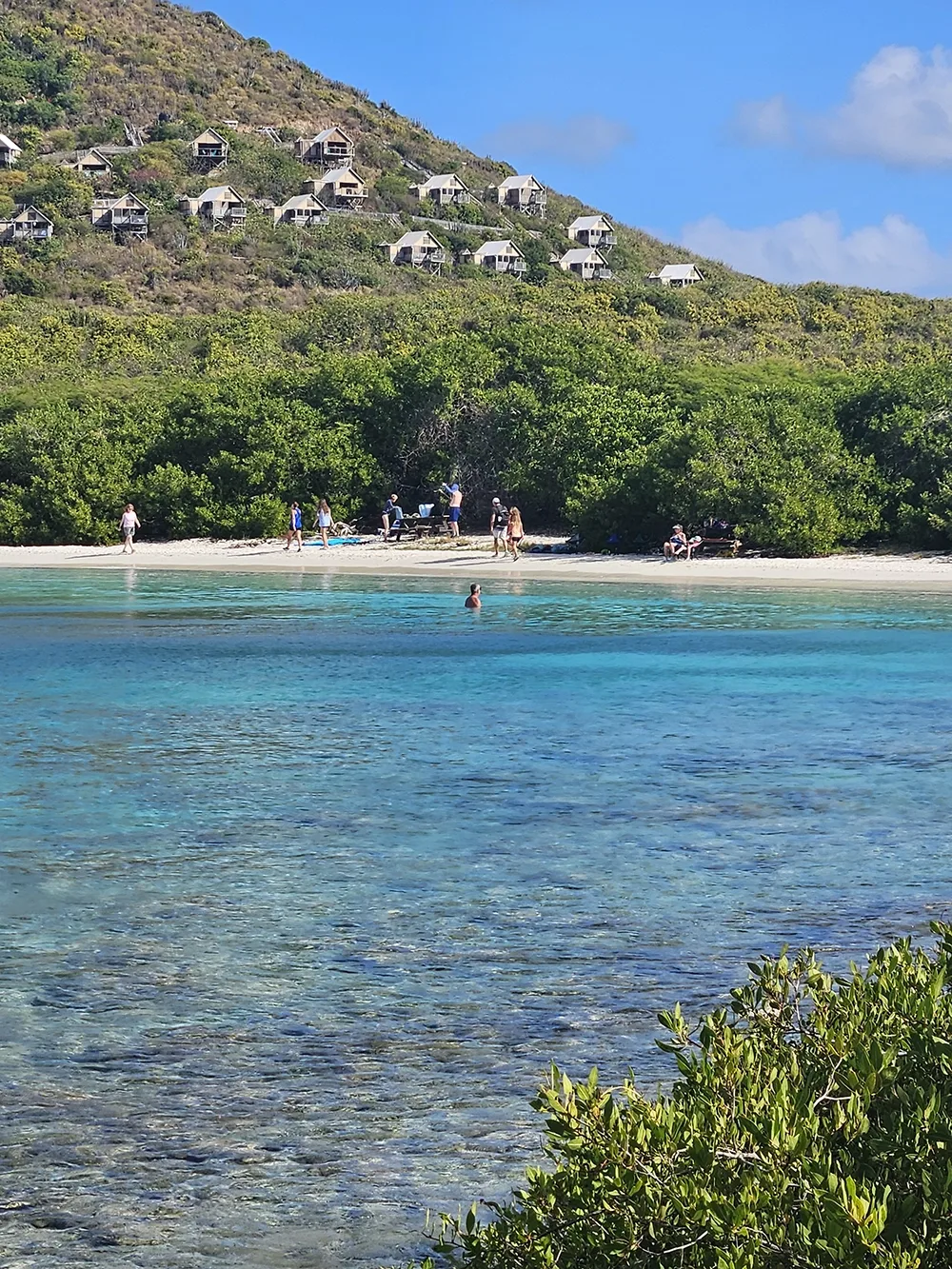 looking across shallow water to a beach area and cabins on the hills behind it