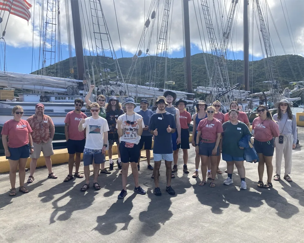 A photo of 18 people standing on a concrete dock in front of a sailing ship with masts, sails, and rigging, with tropical hills in the background.