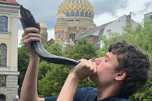 Person blowing a shorar with with a gilded, dome building in the background.