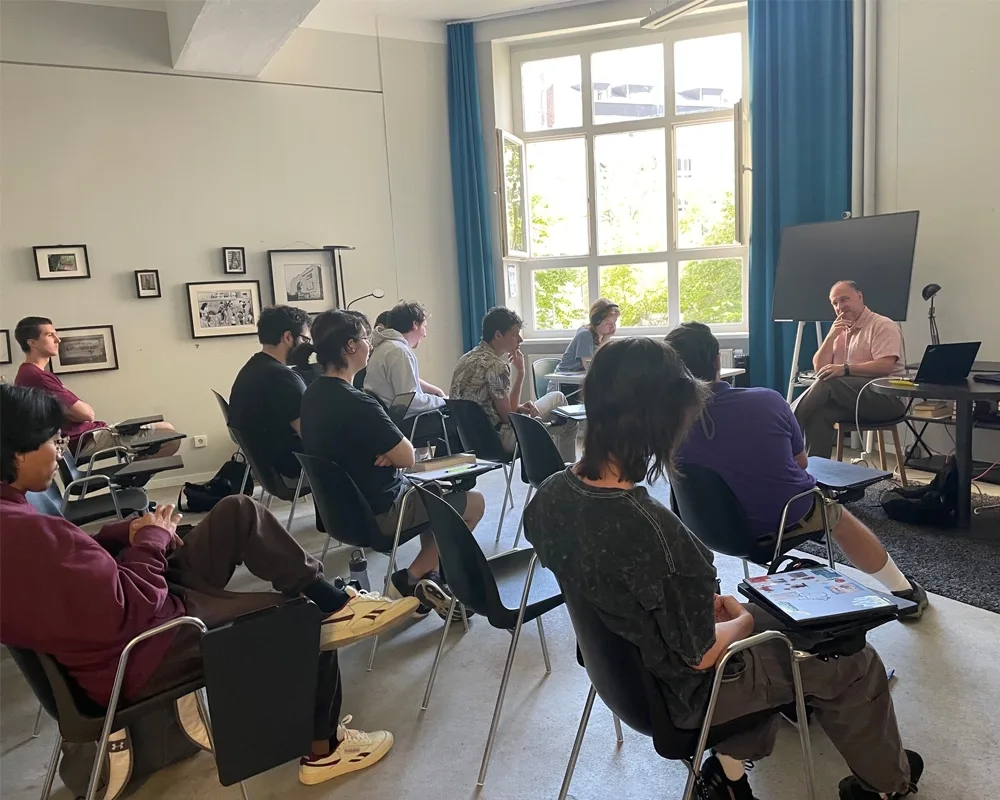 Students sitting in a classroom facing the instructor with a window open in the background.