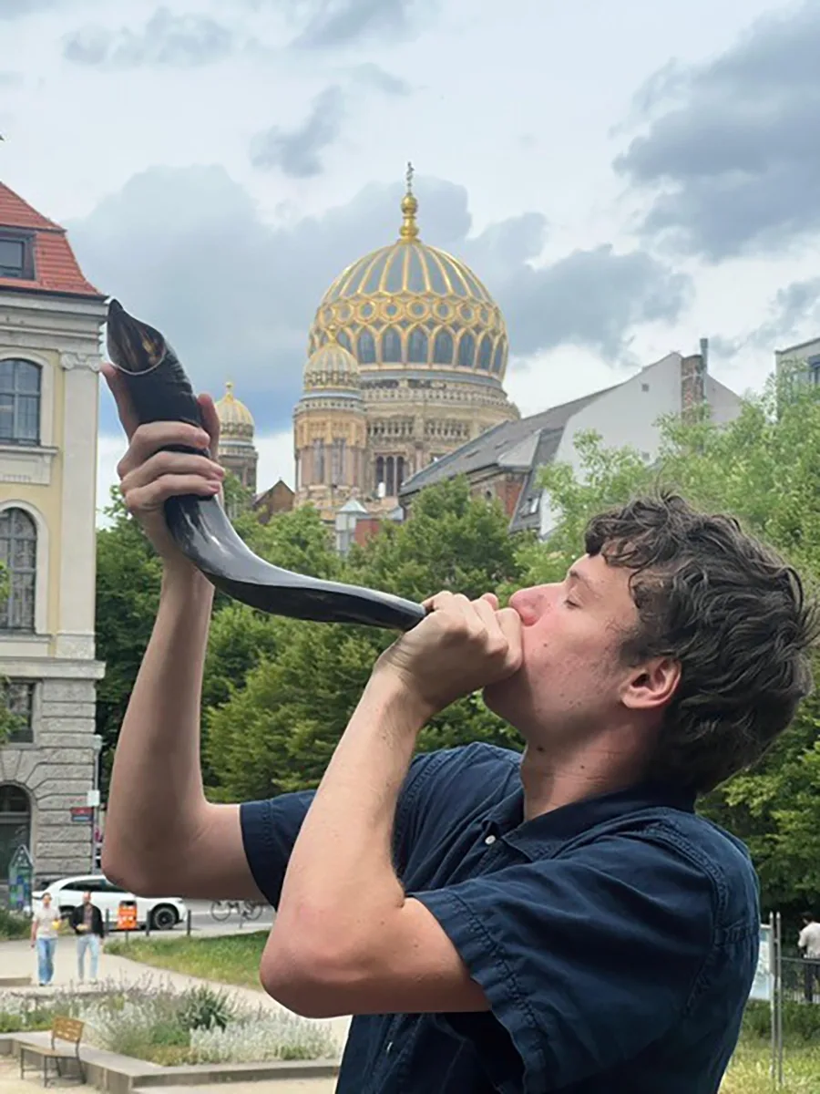 Person blowing a shorar with with a gilded, dome building in the background.