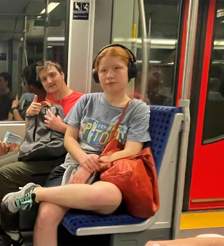 Two students (one with headphones on the other giving a thumbs-up) sitting on a subway car.