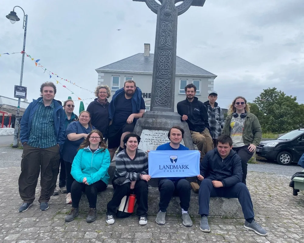 Ireland Program Students in front of a Celtic Cross one holding a small banner that says Landmark College