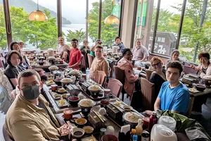 A large group sitting at multiple tables gathers for a group meal with a lake and mountains in the distance.