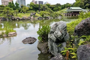 A Japanese garden featuring a pond, teahouse, and urban buildings in the background.