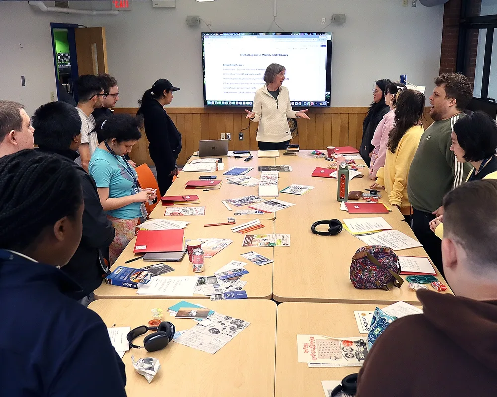 A photo of a professor speaking to a group of students while they are standing around a table.