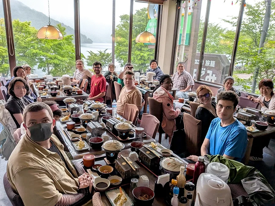 A large group sitting at multiple tables gathers for a group meal with a lake and mountains in the distance.