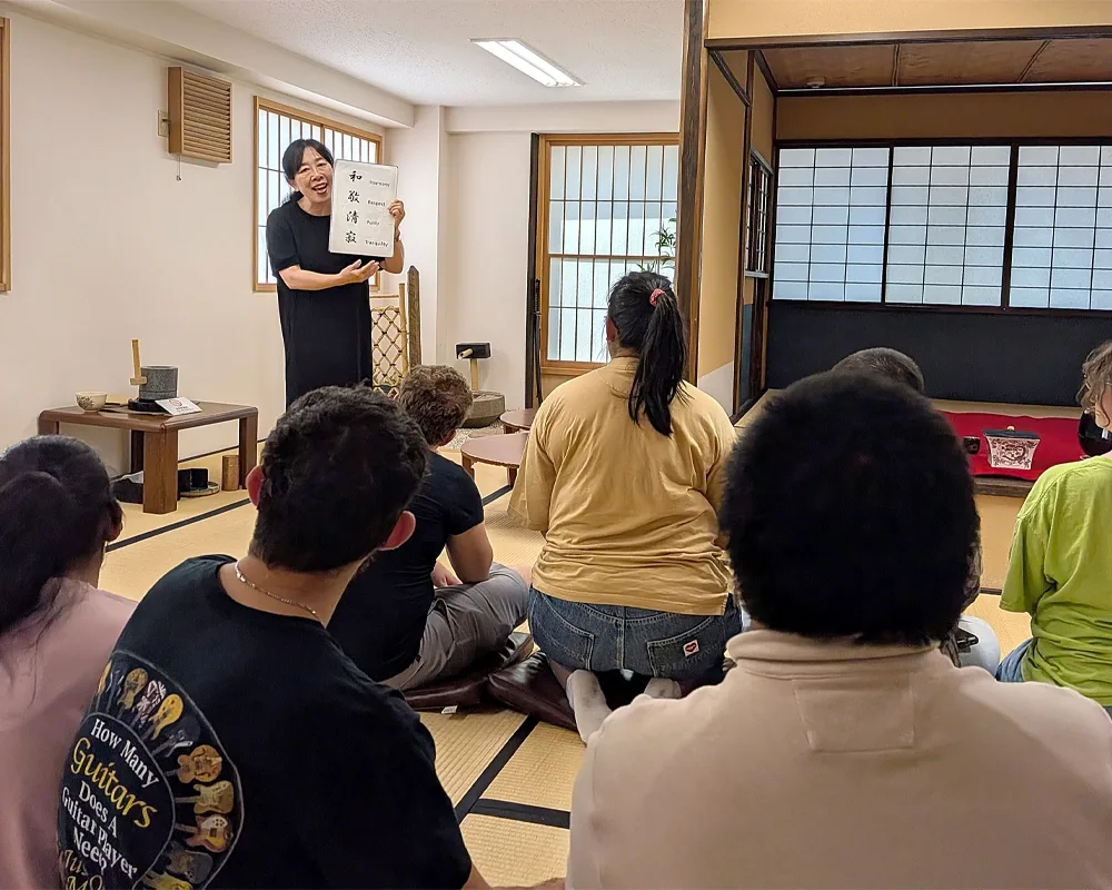A woman speaks to a group while holds a sign featuring Japanese characters for harmony, respect, purity, and tranquility.