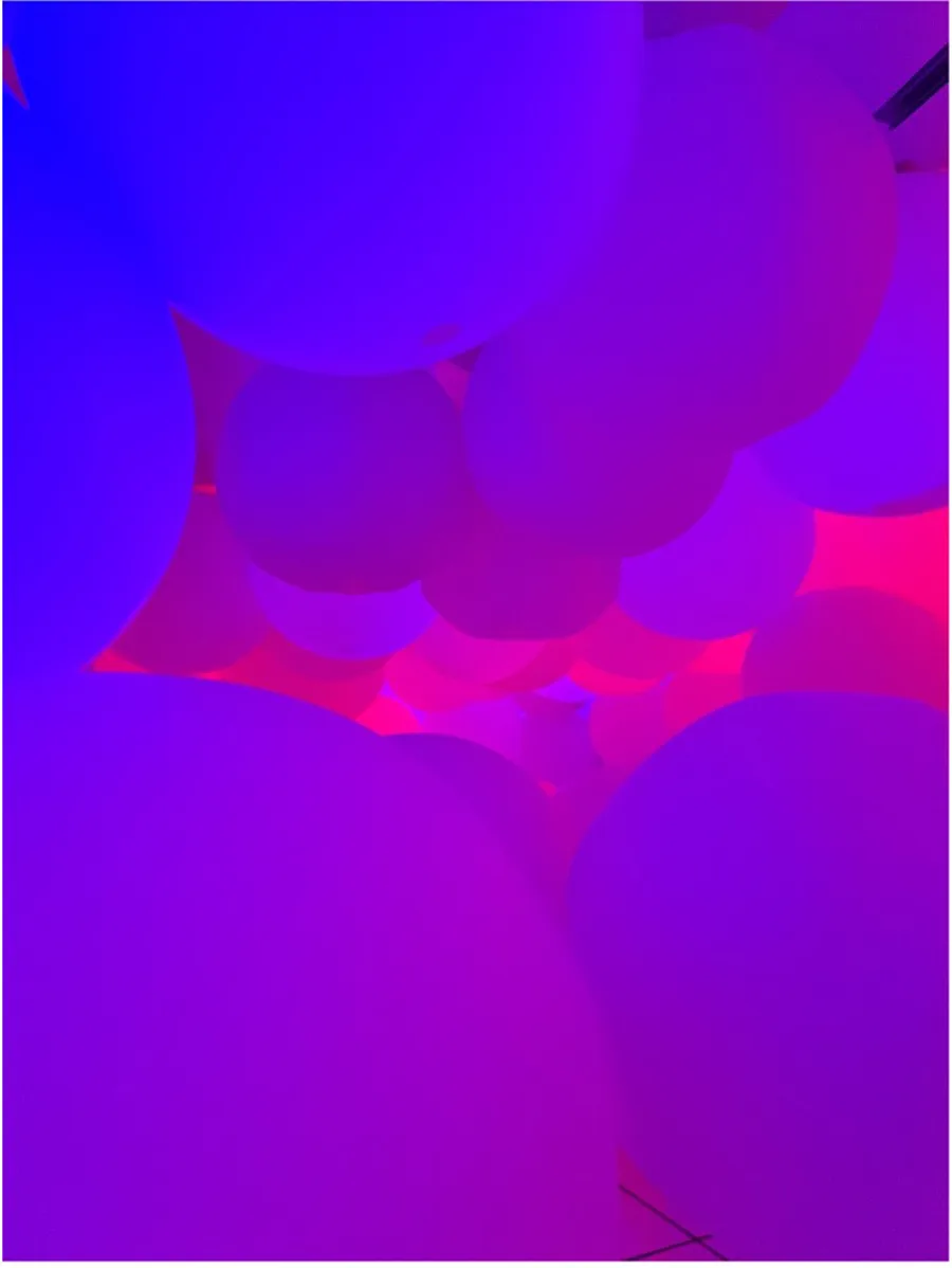 A room filled with large balls light up with blue, pink, and purple lights. The balls are on the floor and hang from the ceiling stacked on top of each other.