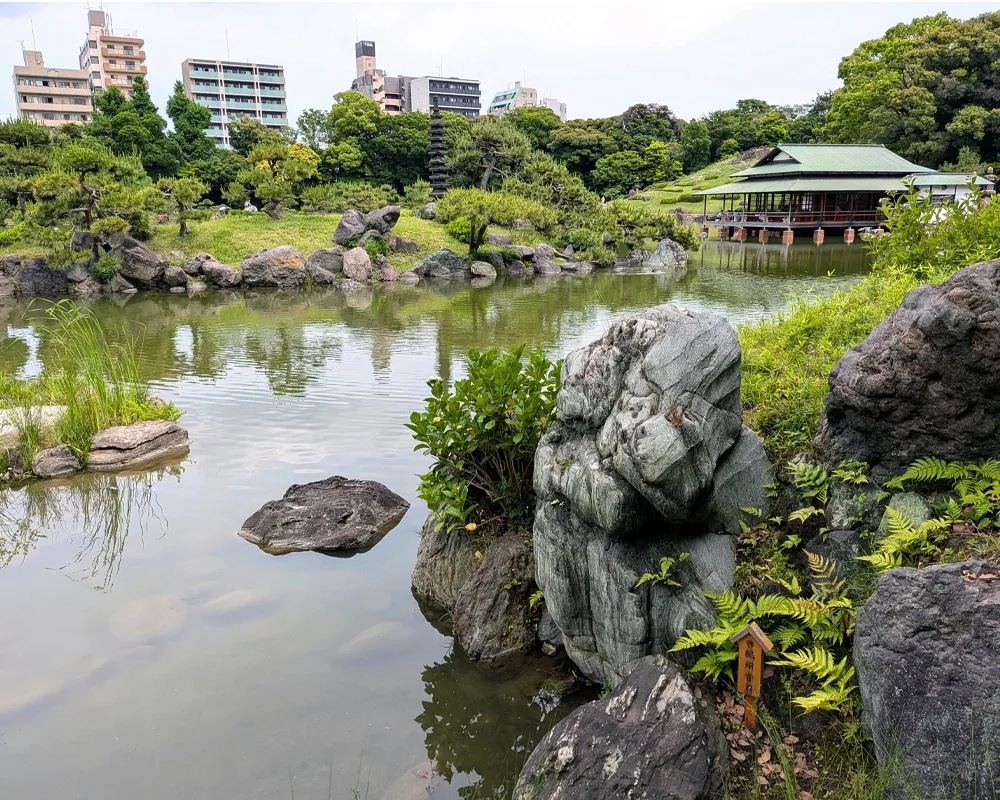 A Japanese garden featuring a pond, teahouse, and urban buildings in the background.