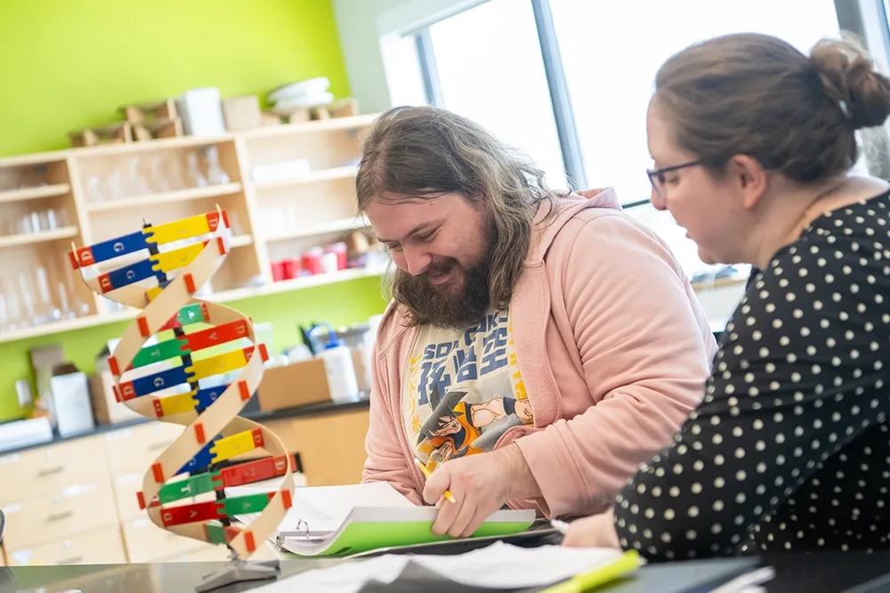 A professor sitting with a student in a lime green room and model of a double helix DNA model in front of them.